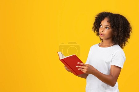 Téléchargez les photos : Positif adolescent surprise élève noire en t-shirt blanc lit le livre, regarde l'espace vide, isolé sur fond jaune, studio. Étudier à l'école, publicité et offre, éducation, mode de vie, passe-temps - en image libre de droit