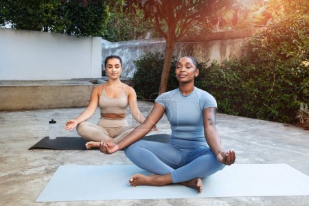Photo for Calm young arab and black women athletes in sportswear practice yoga, meditate in lotus position with closed eyes on mat with bottle of water, outdoor. Fit lifestyle, rest, workout together - Royalty Free Image