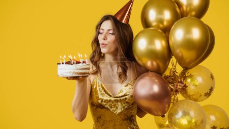 Photo for Pretty european lady in hat with many balloons blowing out candles on cake and making wish, standing isolated on yellow background, panorama. Celebration birthday, holiday party, ad and offer - Royalty Free Image
