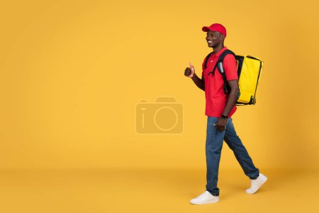 Photo for Positive young african american delivery man in uniform and cap with big backpack go, show thumb up, isolated on orange studio background, full length. Recommendation delivery service, ad and offer - Royalty Free Image