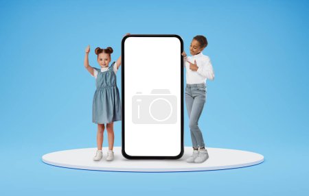 Photo for Two Little Girls Standing Near Huge Blank Smartphone With White Screen, Happy Kids Advertising Mobile App Or Website, Recommending Online Offer, Posing Over Blue Background, Collage, Mockup - Royalty Free Image
