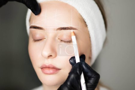 Photo for Professional Cosmetologist Mapping Eyebrows Of Young Woman With White Pencil Before Permanent Makeup Treatment In Beauty Salon, Marking Up Brow Zone And Forehead Before PMU Procedure, Closeup Shot - Royalty Free Image
