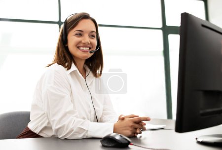 Foto de Comunicación de soporte al cliente. Operadora de línea directa sonriente Mujer de negocios en auriculares que realiza videollamadas en la computadora en el lugar de trabajo, en el interior de la oficina moderna, comunicando y ayudando a los clientes en línea - Imagen libre de derechos