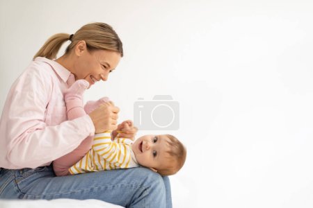 Photo for Happy loving european mother playing with baby daughter, taking care of child, enjoying tender moment on bed, side view, free space. Love mother and kid at home - Royalty Free Image