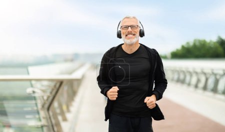 Photo for Positive retired grey-haired sportsman wearing black sportswear and wireless headphones jogging outdoor by bridge in the city, listening to music, smiling. Senior man enjoying workout, blank space - Royalty Free Image