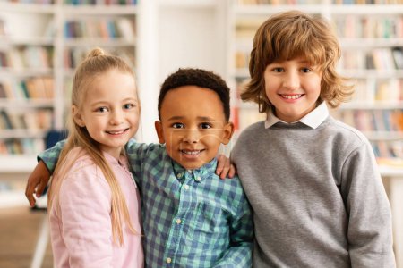 Téléchargez les photos : Portrait de camarades de classe multiraciaux heureux de l'école primaire embrassant et souriant à la caméra, debout dans la salle de classe ou la bibliothèque. Temps scolaire, concept d'éducation et d'amitié - en image libre de droit