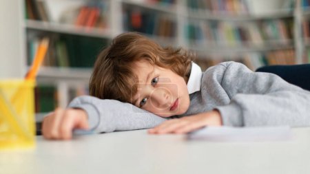 Téléchargez les photos : Petit écolier européen ennuyé regardant ailleurs et pensant tout en s'appuyant sur une table dans une bibliothèque ou une salle de classe pendant une leçon désintéressée, panorama, espace libre - en image libre de droit
