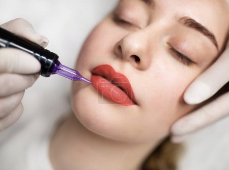 Photo for Lip Blushing. Cosmetologist applying permanent make up on lips of beautiful woman, closeup of attractive young female getting pmu treatment in beauty studio, artist using tattoo pen machine - Royalty Free Image