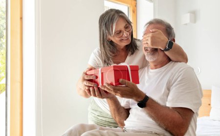 Photo for Happy old european wife closes eyes to her husband, gives gift box in living room interior. Celebration birthday, anniversary, holiday together, surprise and congratulate at home - Royalty Free Image