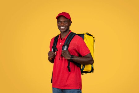 Photo for Happy young african american deliveryman in red uniform with big backpack, enjoy work, isolated on orange studio background. Delivery service, ad and offer, food order at home - Royalty Free Image
