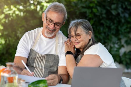 Photo for Smiling senior european wife and husband in aprons make notes at table with laptop, vegetables, enjoy spare time, outdoor. Ordering healthy food at home, new recipe for cooking - Royalty Free Image
