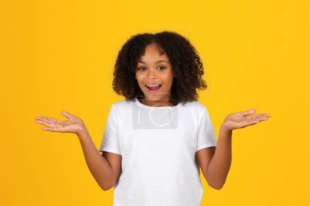 Photo for Glad shocked adolescent black girl in white t-shirt with open mouth spreads arms to sides, holds empty space, isolated on orange background. Choice, surprise, sale, study at school, ad and offer - Royalty Free Image