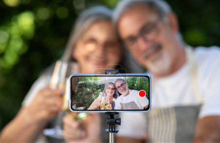 Photo for Happy old european husband and wife take selfie, shoot video blog on smartphone screen, cheers with glasses of wine, outdoor. Cooking food together, device for video call with technology, holiday - Royalty Free Image