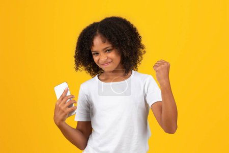 Téléchargez les photos : Joyeux adolescent afro-américaine en t-shirt blanc, utilise le téléphone célébrer le succès, isolé sur fond orange, studio. Gagner, application pour l'étude, la connaissance, l'éducation moderne à l'école, annonce - en image libre de droit