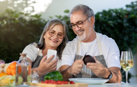 Photo for Cheerful old european couple in aprons with glasses of wine use smartphones, chatting in garden, outdoors. Lunch holiday together, device for video call, technology and fun - Royalty Free Image