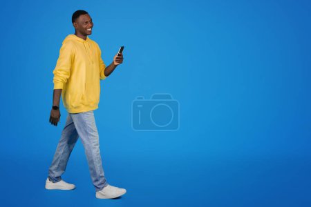 Photo for Cheerful young african american guy in hoodie uses smartphone, isolated on blue studio background, full length. Free time, chat, social media app and lifestyle, ad and offer - Royalty Free Image