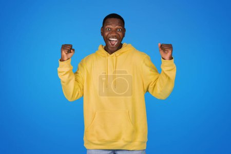 Photo for Cheerful surprised excited young african american man in sweatshirt raises hands up, celebrating victory, isolated on blue studio background. Success in study, work, win, good news ad and offer - Royalty Free Image