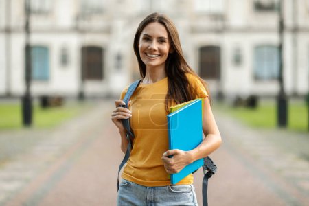 Smiling Young Female Student With Workbooks And Backpack Standing Outdoors, Happy Beautiful Millennial Woman Posing Outside At University Campus, Holding Books And Looking At Camera, Copy Space