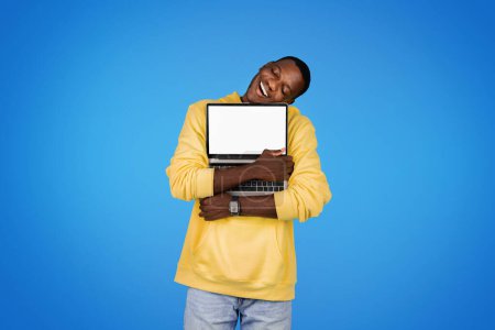 Téléchargez les photos : Joyeux jeune homme afro-américain en sweat-shirt étreignant ordinateur portable avec écran blanc, isolé sur fond de studio bleu. Fun, technologie moderne et style de vie, annonce et offre le travail et l'étude de l'amour - en image libre de droit