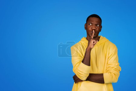 Photo for Pensive young african american guy in hoodie presses finger to lips, making shh sign, isolated on blue studio background. Lifestyle, secret and surprise, idea creation, ad and offer - Royalty Free Image