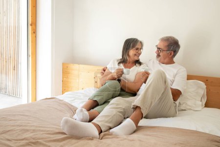 Photo for Cheerful old european man and lady have breakfast with cups of coffee, enjoy free time, sit on bed in bedroom interior. Communication and relationships in morning at home, rest and relax together - Royalty Free Image