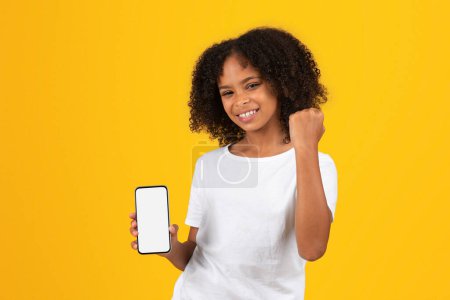 Photo for Glad teenage black pupil girl in white t-shirt shows phone with blank screen, makes success, victory sign with hand, isolated on yellow background, studio. Study, good news and school, ad and offer - Royalty Free Image