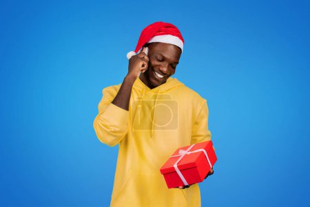 Photo for Cheerful young african american guy in sweatshirt and Santa hat, calls by phone, show gift box, isolated on blue studio background. Communication, great news, present for New Year, Christmas - Royalty Free Image