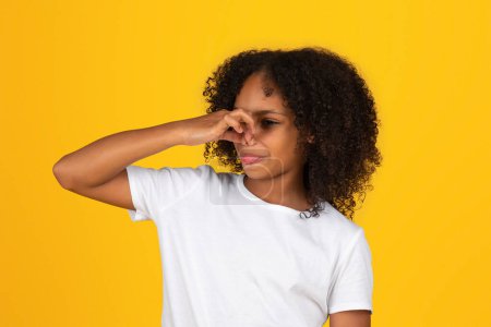 Photo for Sad adolescent african american girl in white t-shirt covering nose, suffering from bad smell, isolated on orange background, studio. Stink, disgust, problems with hygiene, human emotions, study - Royalty Free Image