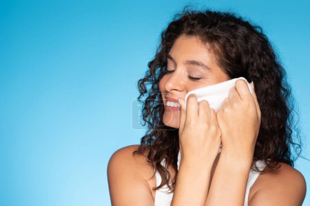 Photo for Cheerful young european curly woman enjoy skin moisturizing, cleanse face with towel, isolated on blue studio background. Spa and daily treatments, beauty care and cosmetics remove - Royalty Free Image