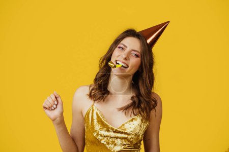 Photo for Gorgeous lady in elegant dress and birthday cap blowing party horn, celebrating holiday on yellow studio background. Happy woman having party. Festive concept - Royalty Free Image