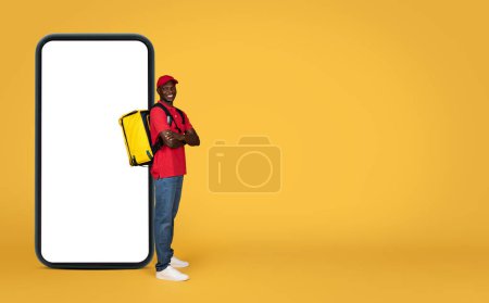 Photo for Happy confident young black delivery man in uniform with big backpack with crossed arms near phone with blank screen, isolated on orange background. Website, delivery service, recommendation app - Royalty Free Image