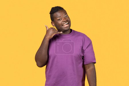 Téléchargez les photos : Homme afro-américain joyeux geste Call Me, tenant la main comme un téléphone près de l'oreille posant sur fond jaune, portant un t-shirt violet. Contact, numéro de téléphone et concept de communication - en image libre de droit