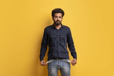 Photo for Poverty Concept. Portrait Of Upset Young Indian Man Showing His Empty Pockets And Looking At Camera, Broke Poor Millennial Guy Posing Over Yellow Background, Suffering Financial Crisis, Copy Space - Royalty Free Image