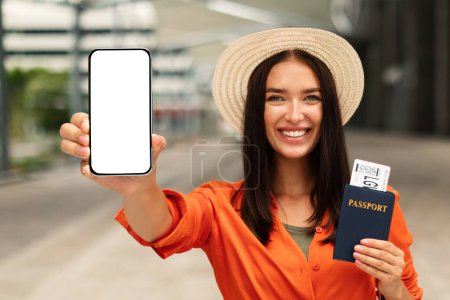 Foto de Alegre señora pasajero mostrando Smartphone pantalla vacía y mostrando tarjeta de embarque en el pasaporte, recomendando la aplicación móvil para la reserva de entradas y planificación de viajes al aire libre, burla - Imagen libre de derechos