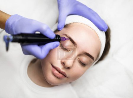 Photo for Beautician hand doing eyebrow permanent makeup on young attractive female face, professional pmu artist in gloves making microblading tattoo to brows of beautiful woman, using machine pen, closeup - Royalty Free Image