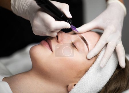 Photo for Cosmetologist applying permanent makeup on eyebrows of beautiful young female, calm attractive woman getting eyebrow tattoo by professional artist, lying on table with closed eyes, enjoying procedure - Royalty Free Image