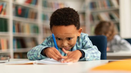 Photo for Concentrated black schoolboy sitting at desk in school and learning to write in copybook with pencil, studying, panorama. Diverse children in the class - Royalty Free Image