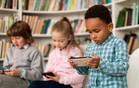 Téléchargez les photos : Divers élèves de l'école utilisant des smartphones dans la bibliothèque, des enfants multiraciaux tenant des téléphones mobiles, jouant à des jeux ou regardant des vidéos en ligne pendant la pause scolaire - en image libre de droit