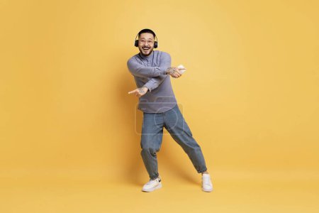 Foto de Tiro de longitud completa de alegre joven asiático bailando sobre fondo amarillo, Feliz Millennial Guy usando auriculares inalámbricos y sosteniendo un teléfono inteligente moderno en la mano, Disfrutando de canciones favoritas, Copiar el espacio - Imagen libre de derechos
