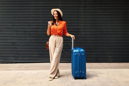 Téléchargez les photos : App de planification de voyage. Joyeuse voyageuse avec valise utilisant l'application téléphonique pour réserver des billets, debout à l'extérieur près du mur de l'aéroport, portant un chapeau et des vêtements décontractés. Vacances avec la technologie mobile - en image libre de droit