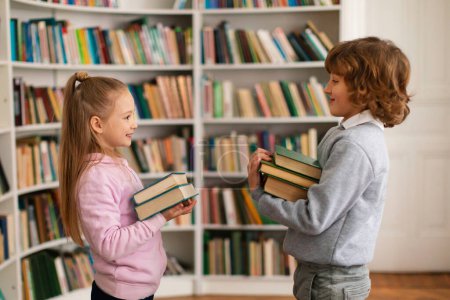 Photo for School boy and girl holding stack of books and talking with each other standing in library, side view. Happy two children having a conversation, discussing study in primary school - Royalty Free Image