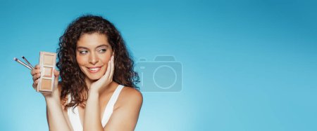 Photo for Cheerful young european curly woman enjoy makeup, touching face with hand, show powder, blush pallet and brushes, isolated on blue studio background. Beauty care and cosmetics recommendation - Royalty Free Image