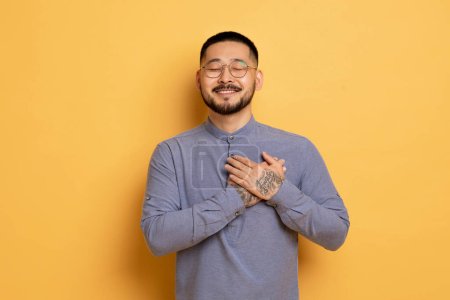 Photo for Gratitude Concept. Thankful Young Asian Man Keeping Both Palms On Chest, Smiling Millennial Male Expressing Appreciation And Kindness While Standing Over Yellow Studio Background, Copy Space - Royalty Free Image