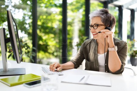 Photo for Cheerful attractive short-haired mature lady sitting at workdesk at home office, using computer pc and headset, have video conference with colleagues or business partner. Woman working at call-centre - Royalty Free Image