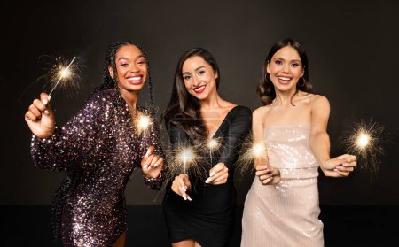 Foto de Loco tiempo de fiesta de tres hermosas mujeres elegantes en traje elegante celebrando año nuevo 2024 juntos sobre fondo negro, damas jóvenes divirtiéndose, bailando, sosteniendo luces de bengala y sonriendo - Imagen libre de derechos