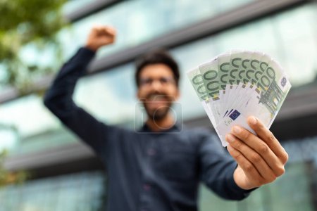 Photo for Excited middle eastern businessman holding money cash, recommending investment, trading and cashback, standing outdoors next to office building, selective focus on euro banknotes, copy space - Royalty Free Image