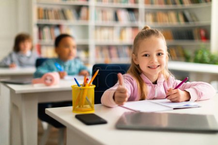Photo for Pretty smart school girl doing thumbs up gesture while studying and sitting at desk in classroom, ready to study for excellence. Education concept - Royalty Free Image