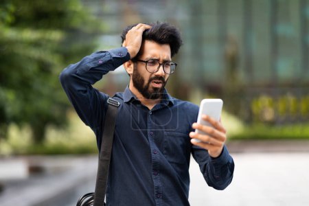 Photo for Upset sad young eastern businessman reading bad news online using app on phone, thinking man outside office building disappointed with achievement results at workplace, boss depressed, touching head - Royalty Free Image