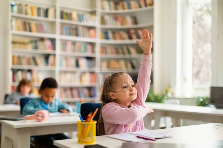 Téléchargez les photos : Étudiante intelligente du primaire levant la main, prête à répondre aux questions des enseignants à l'intérieur de la salle de classe. Fille assise au bureau avec des camarades de classe sur le fond, espace libre - en image libre de droit