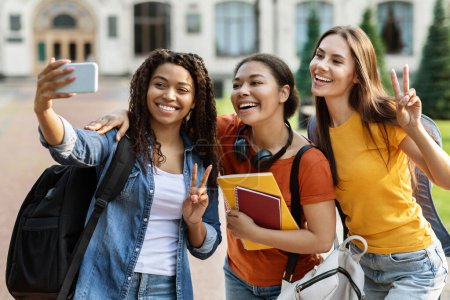 Téléchargez les photos : Trois étudiantes multiethniques parlent selfie sur smartphone en plein air, groupe d'amis de collège avec des sacs à dos et des cahiers de travail posant ensemble à la caméra de téléphone mobile, se détendre après les cours - en image libre de droit